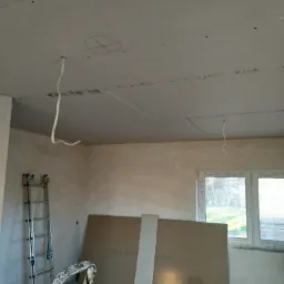 Sufit z płyt kartonowo-gipsowych w trakcie montażu, widoczne kable wystające z otworów, ślady po ołówku, drabina oparta o ścianę, płyty gipsowe oparte o ścianę przy oknie.