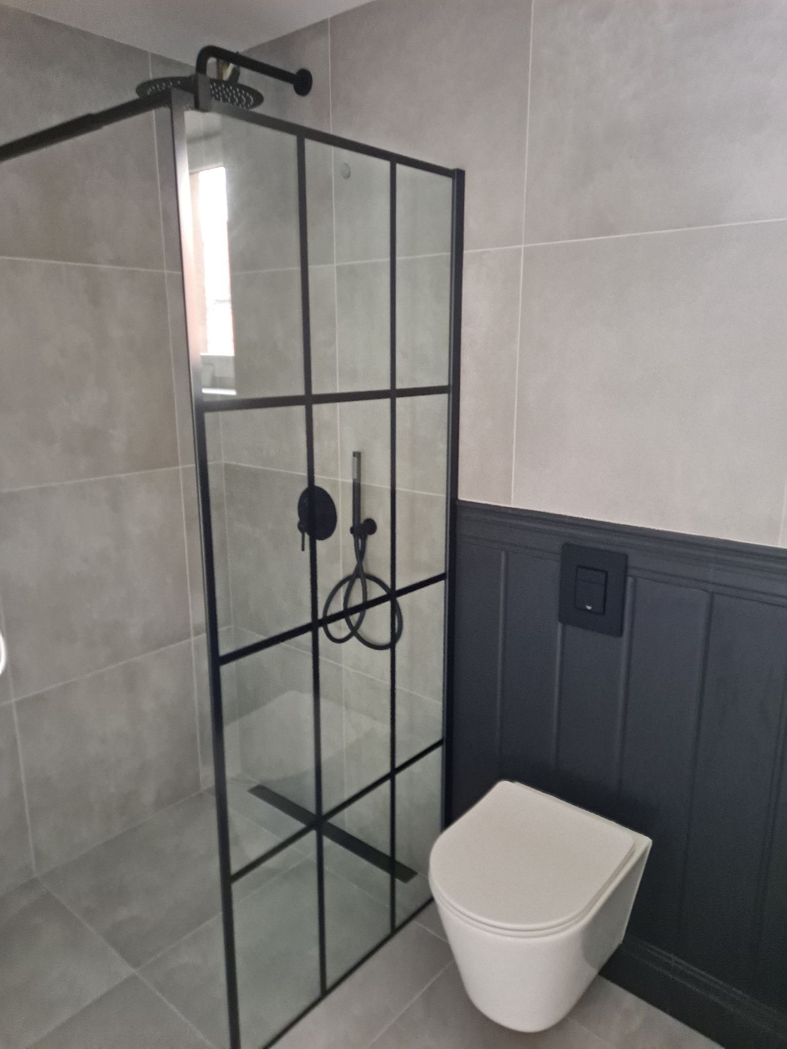 Nowoczesna łazienka z kabiną prysznicową typu walk-in z czarnymi profilami, wiszącą toaletą i szarą płytką na ścianach.