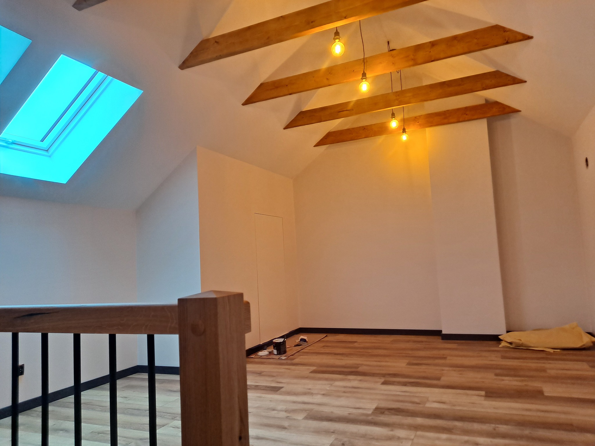 Poddasze po remoncie z drewnianymi belkami stropowymi, trzema żarówkami na kablach, oknem dachowym i panelami podłogowymi. Widoczne puszki farb i wałek do malowania przy ścianie.