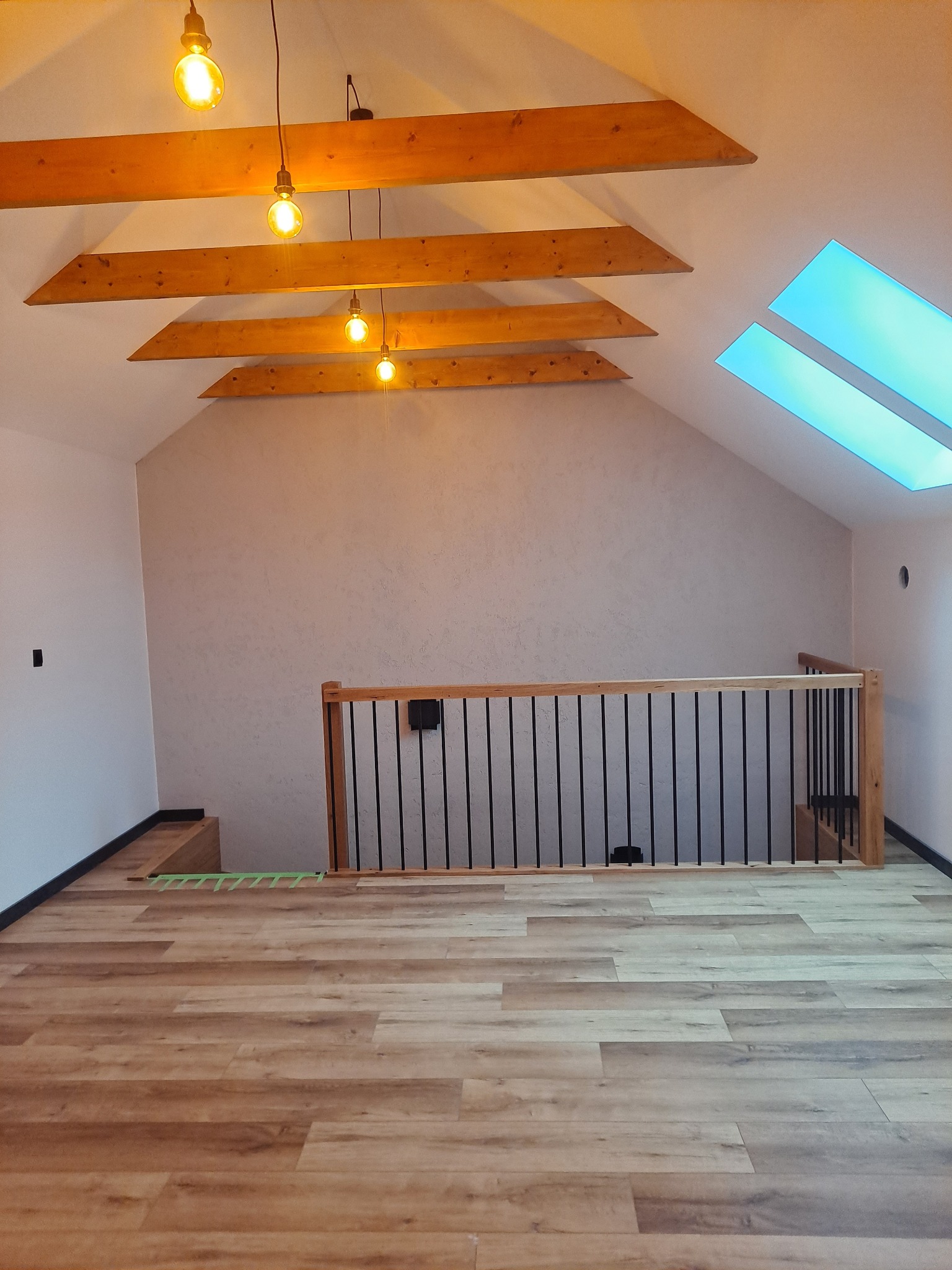 Poddasze z drewnianą balustradą, panelami podłogowymi w odcieniach brązu, drewnianymi belkami stropowymi i oknami dachowymi z niebieskim światłem, oświetlone wiszącymi żarówkami.