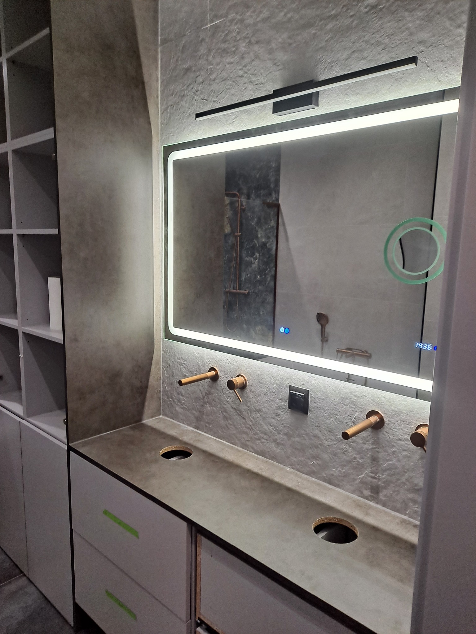 Nowoczesna łazienka z podświetlanym lustrem, armaturą w kolorze miedzi i blatem z otworami na umywalki, widoczne szare ściany o fakturze betonu oraz białe szafki z zielonymi paskami.