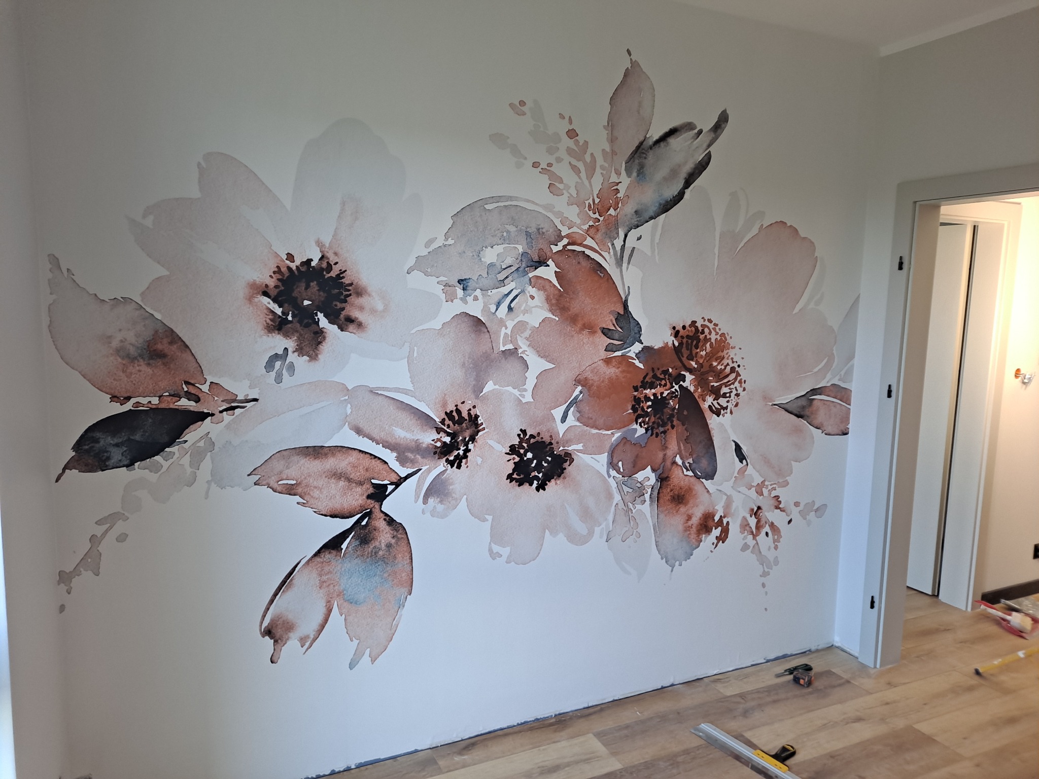 Kwiatowy motyw akwarelowy w odcieniach brązu i beżu na białej ścianie w pokoju, widoczne panele podłogowe i fragment otwartych drzwi do kolejnego pomieszczenia.