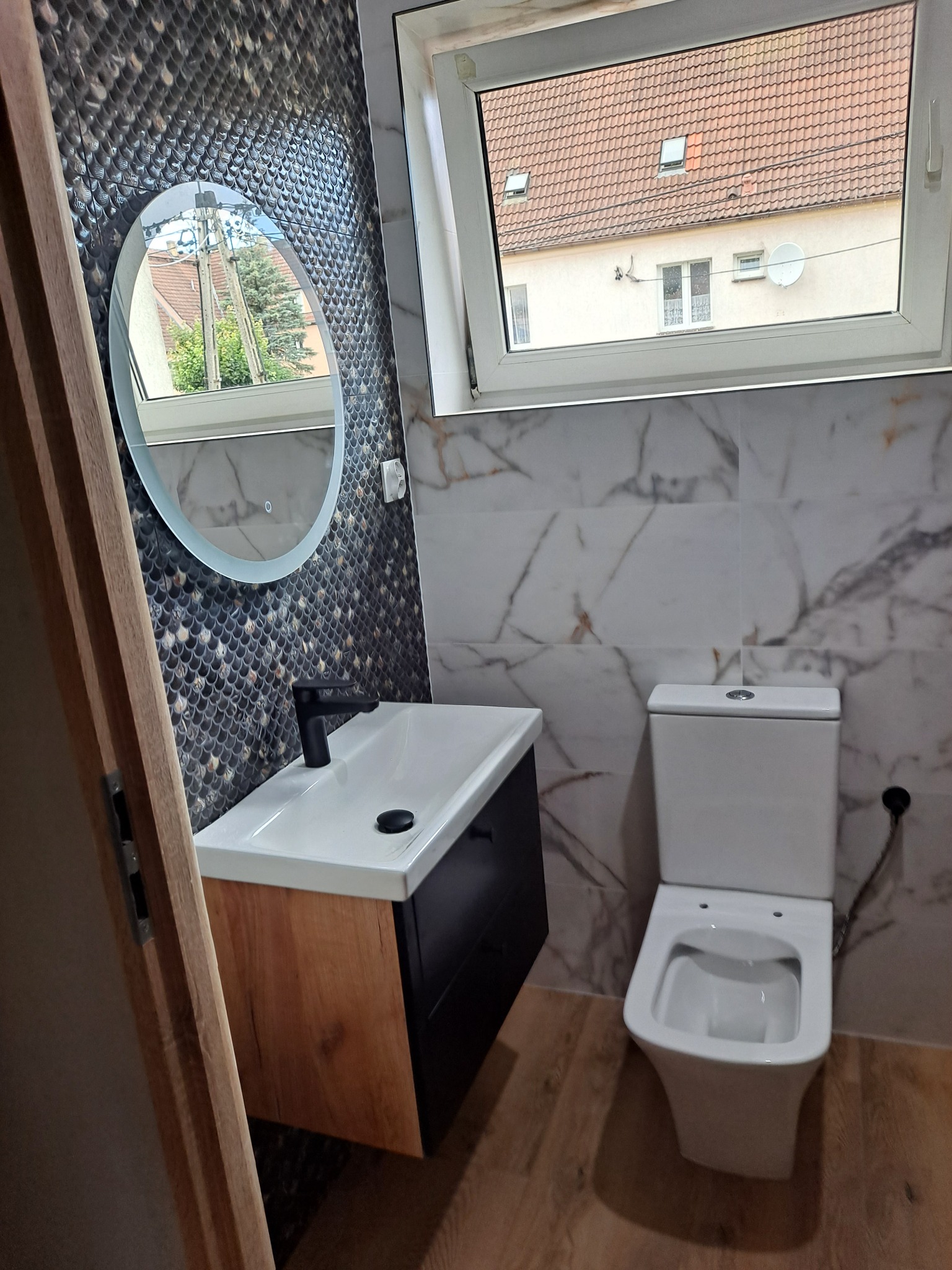 Nowoczesna łazienka po remoncie: biała umywalka z czarną armaturą na szafce z drewnianym frontem, okrągłe lustro z podświetleniem, wisząca toaleta, marmurowe płytki na ścianach i drewnopodobne...