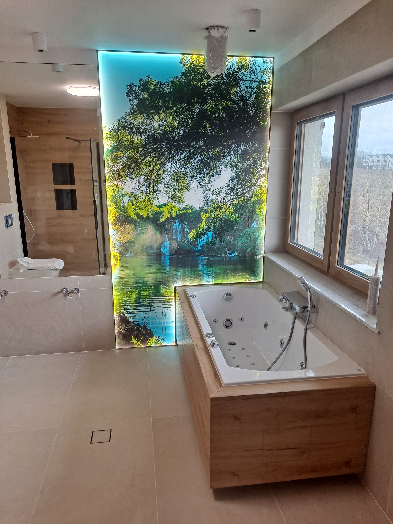 Nowoczesna łazienka z wanną jacuzzi w obudowie z drewna i podświetlanym panelem z motywem wodospadu. Jasne płytki i okno doświetlają wnętrze.