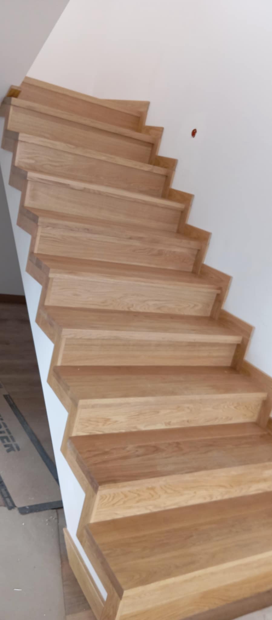 Drewniane schody z jasnego drewna, prowadzące w górę, z widocznym fragmentem białej ściany. Widok z dołu, prezentujący detale wykończenia stopni.