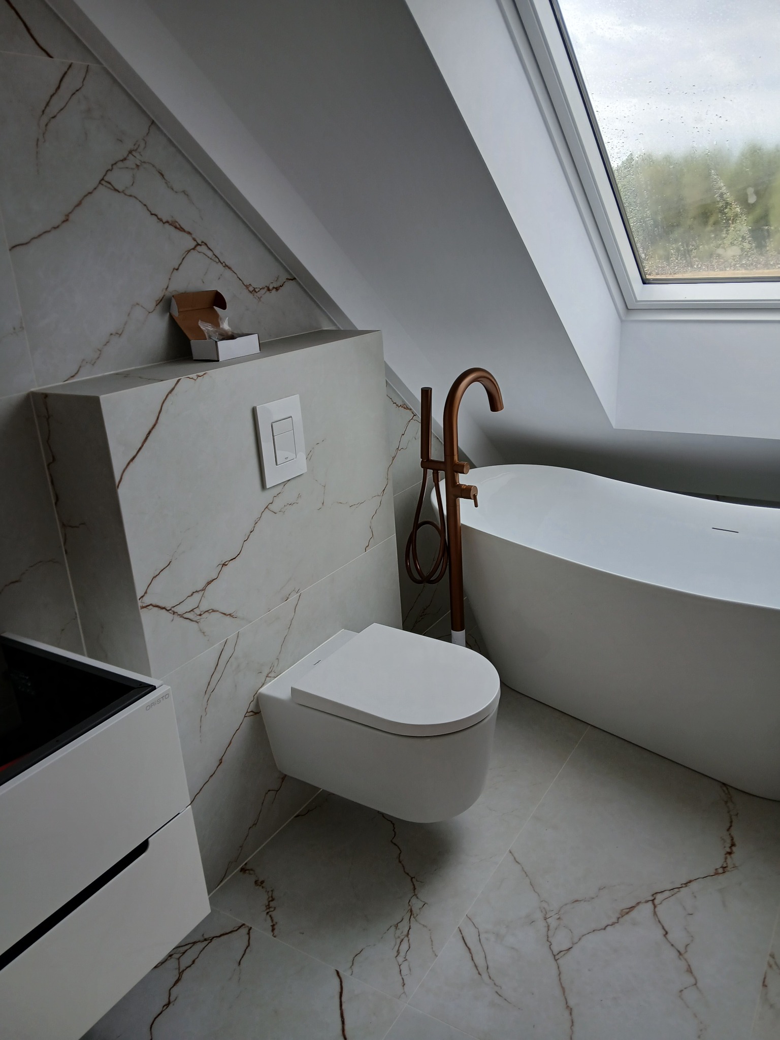 Nowoczesna łazienka na poddaszu z marmurowymi płytkami, białą armaturą i miedzianą baterią wolnostojącą. Na ścianie widoczny przycisk spłuczki i pudełko z akcesoriami.