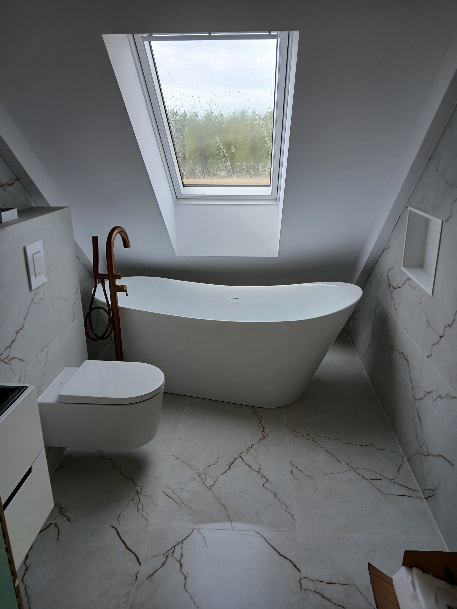 Nowoczesna łazienka na poddaszu z wanną wolnostojącą, toaletą podwieszaną i marmurowymi płytkami. Okno dachowe wpuszcza naturalne światło.