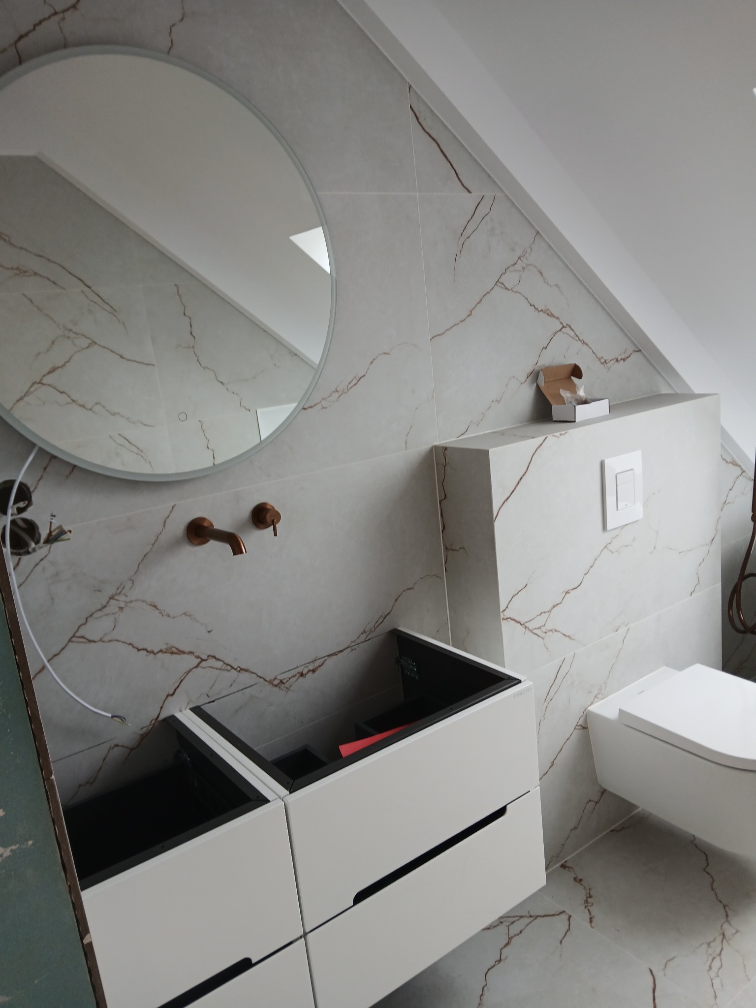 Łazienka na poddaszu w trakcie wykańczania: marmurowe płytki, okrągłe lustro z podświetleniem, biała szafka pod umywalkę i wisząca toaleta. Widoczne elementy instalacji.