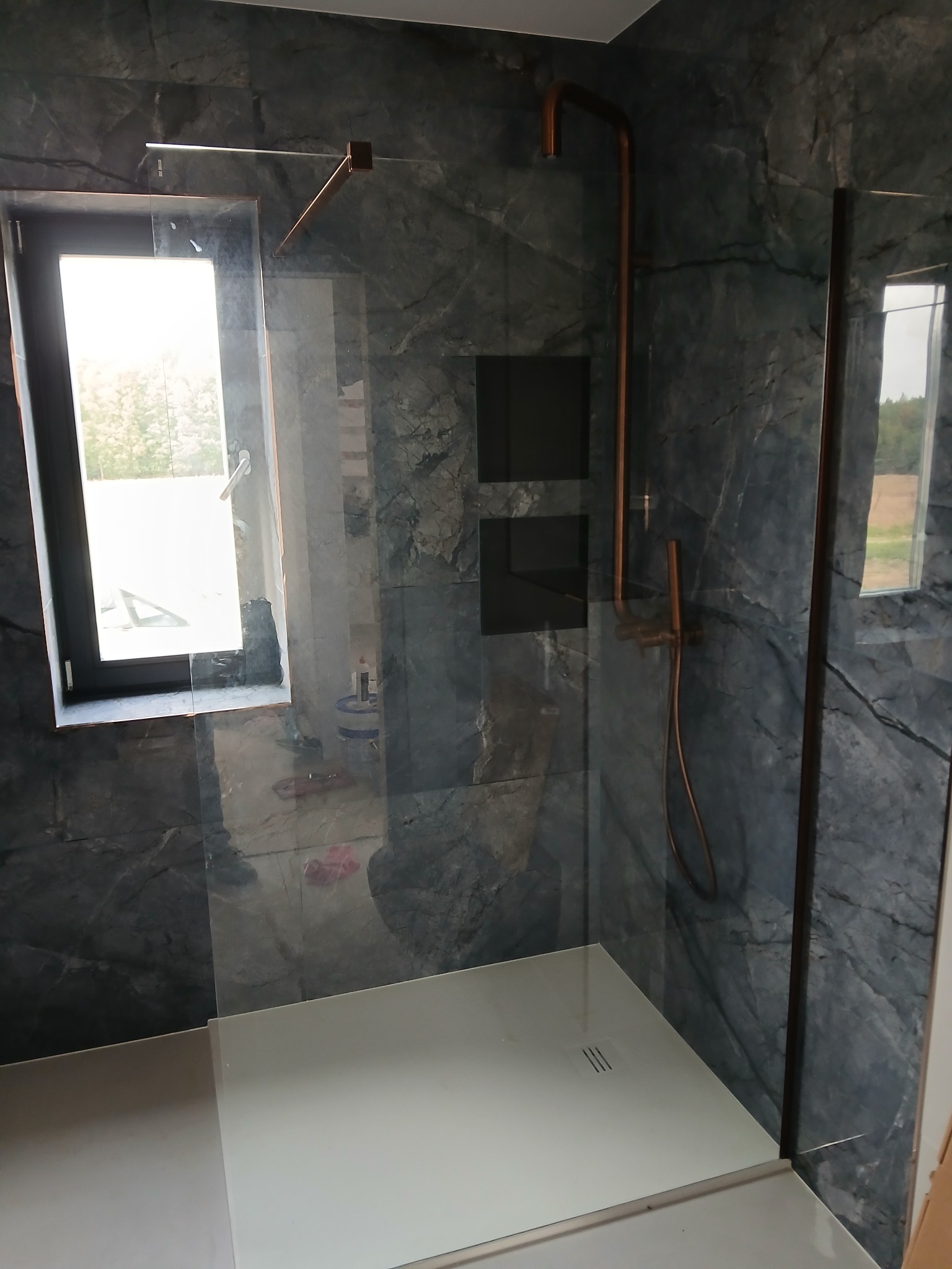 Kabina prysznicowa walk-in z marmurowymi płytkami, miedzianą armaturą i oknem z widokiem na zieleń. Nowoczesny design z dbałością o detale.