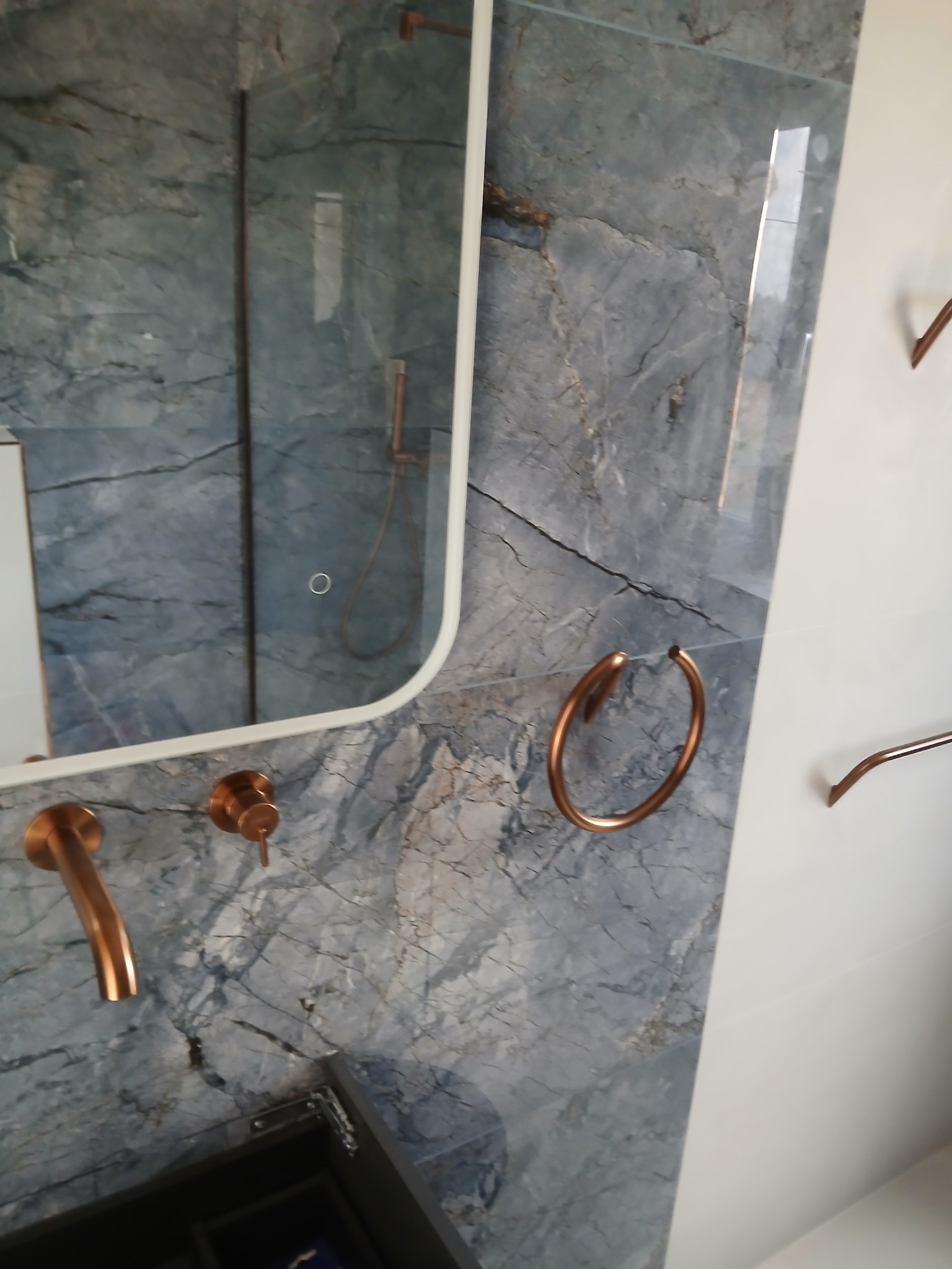 Wykończona łazienka z marmurową ścianą, lustrem i miedzianą armaturą. Nowoczesny design z akcentami miedzi i szarości. Eleganckie wykończenie.