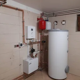 Instalacja kotłowni z białym zasobnikiem ciepłej wody, czerwonym zbiornikiem wyrównawczym i białym modułem sterującym, połączona miedzianymi rurami w pomieszczeniu z beżowymi płytkami.