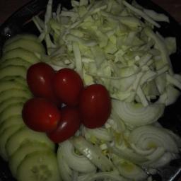 Kuchnia Madziejki - Czarny talerz z przygotowanymi składnikami sałatki: plasterki ogórka, pomidorki koktajlowe, poszatkowana kapusta i cebula.