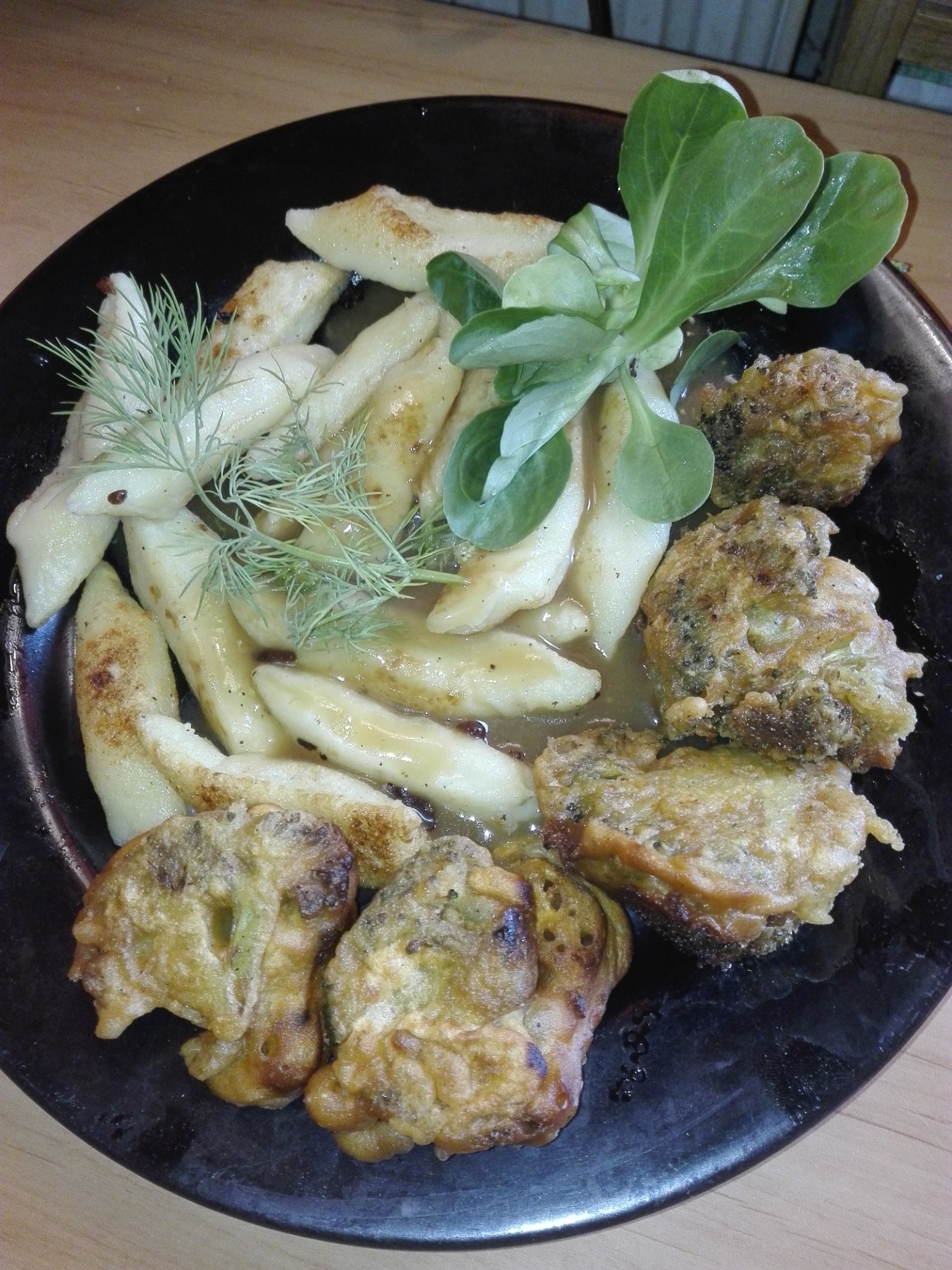 Kopytka polane sosem, podane z brokułami w chrupiącej panierce, udekorowane świeżym koperkiem i roszponką na czarnym talerzu.