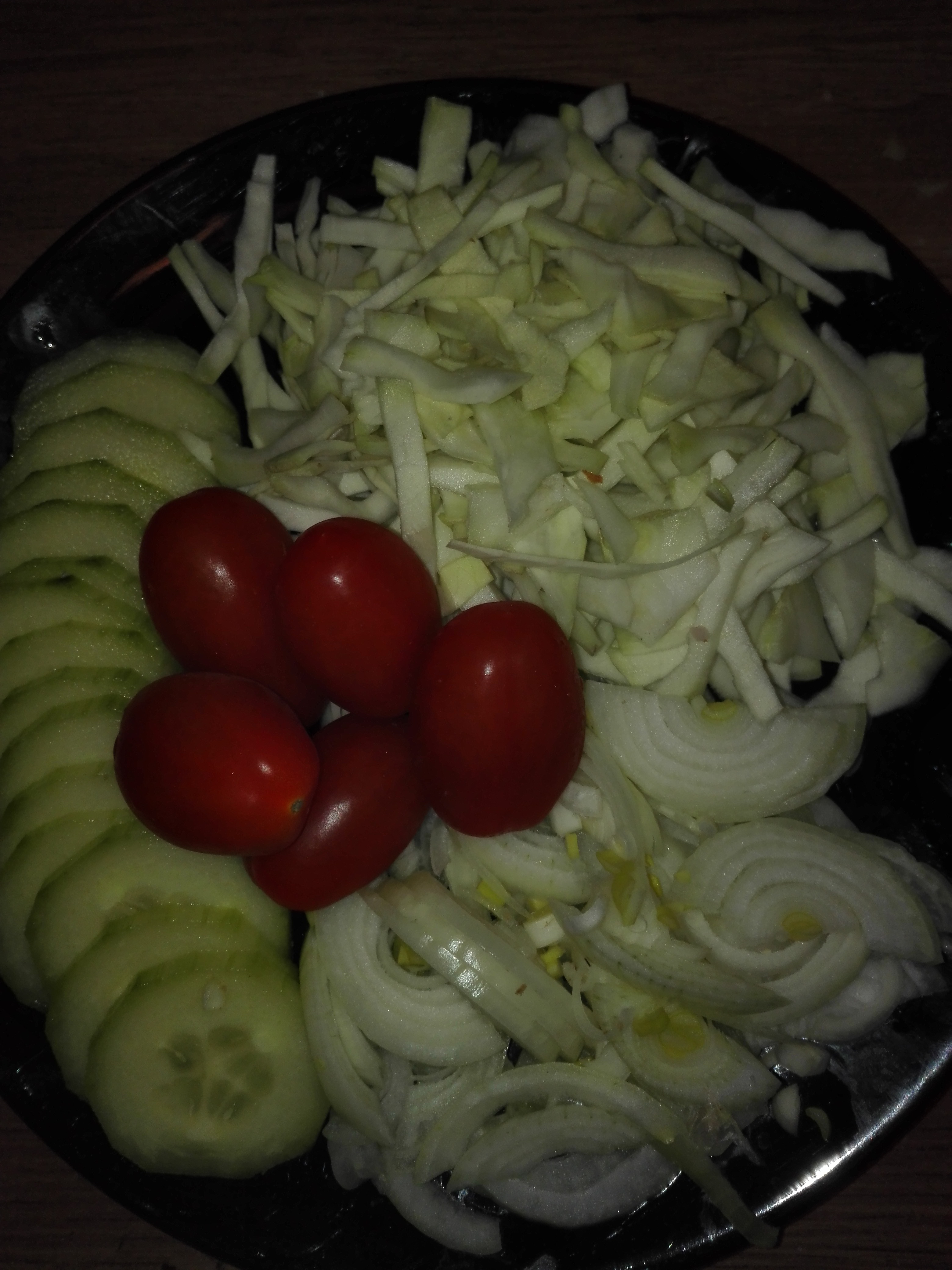 Czarny talerz z przygotowanymi składnikami sałatki: plasterki ogórka, pomidorki koktajlowe, poszatkowana kapusta i cebula.