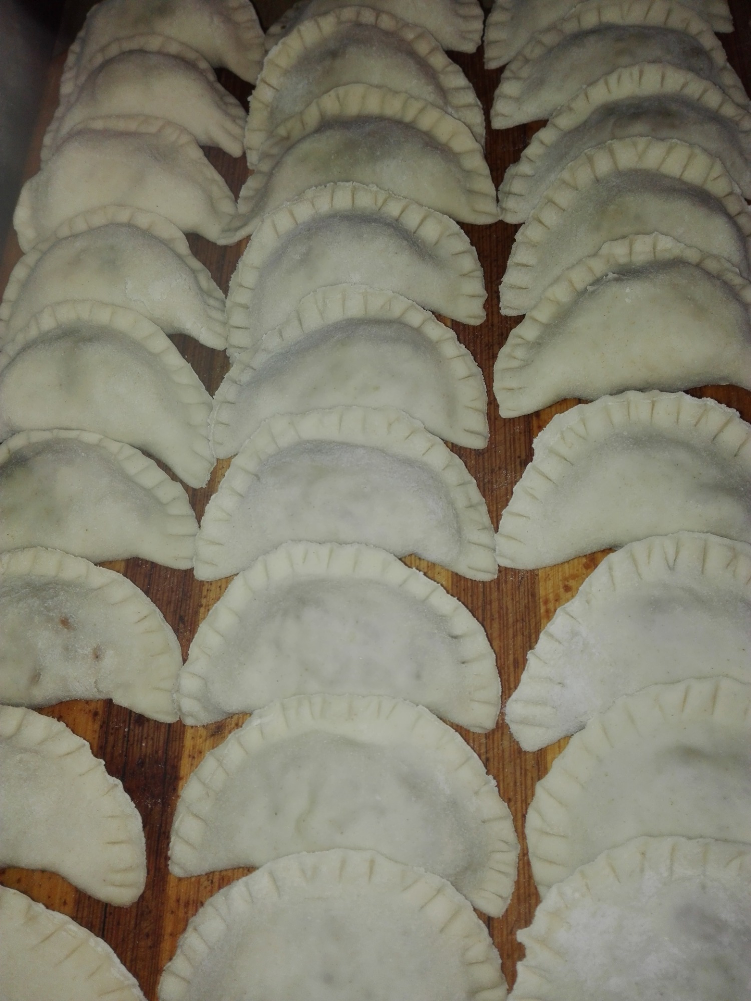 Surowe pierogi z mięsem ułożone rzędami na drewnianej desce, przygotowane do gotowania lub mrożenia, z charakterystycznym karbowanym brzegiem.