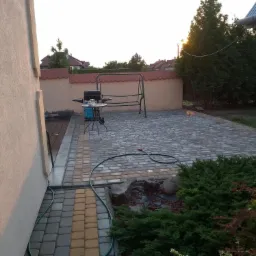 Ułożona kostka brukowa na podwórku z grillem i huśtawką w tle, widoczny wąż ogrodowy i roślinność.