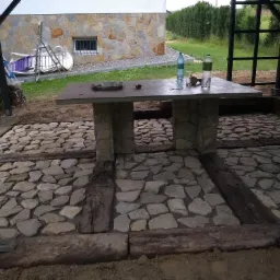 Kamienna podłoga tarasu w trakcie budowy, z drewnianymi belkami i kamiennym stołem w tle domu z elewacją z kamienia naturalnego.