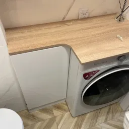 Blat drewnopodobny nad zabudowaną pralką w łazience, widoczny fragment umywalki i dekoracyjne patyczki zapachowe.