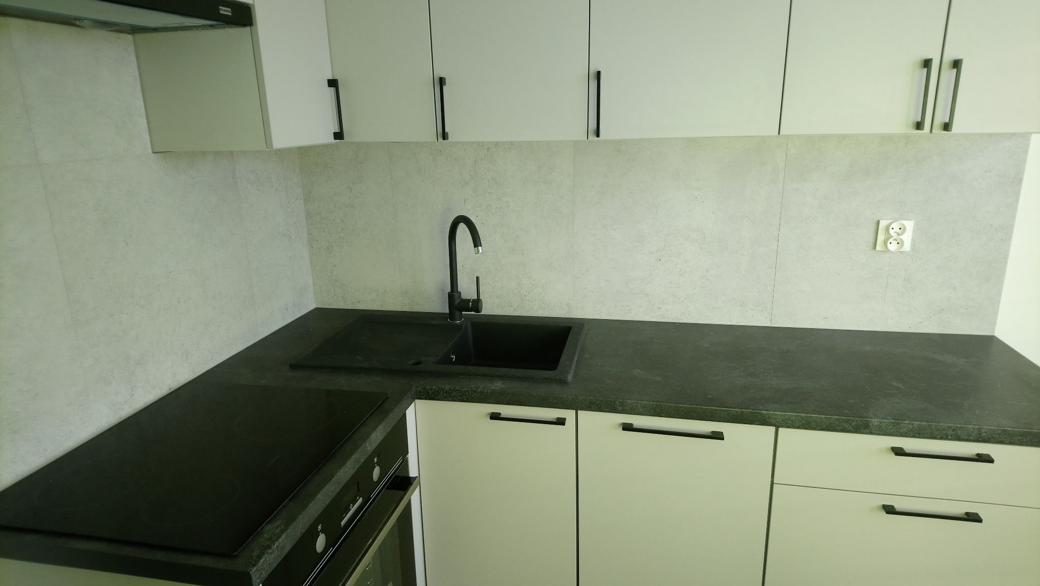 Nowoczesna kuchnia z białymi szafkami, czarnym blatem i zlewozmywakiem, minimalistyczne uchwyty, szara ściana nad blatem z gniazdkiem elektrycznym.