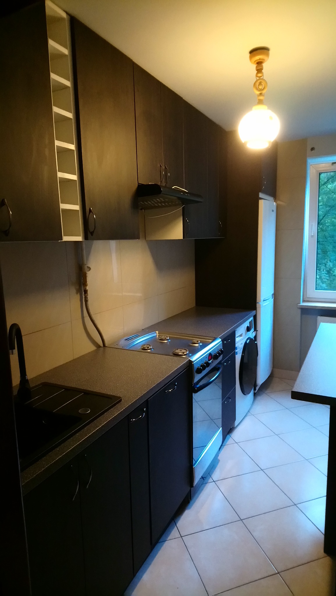 Ciemna, nowoczesna kuchnia z grafitowymi szafkami, jasnymi płytkami na ścianie i podłodze, oraz widoczną kuchenką gazową i pralką pod blatem.