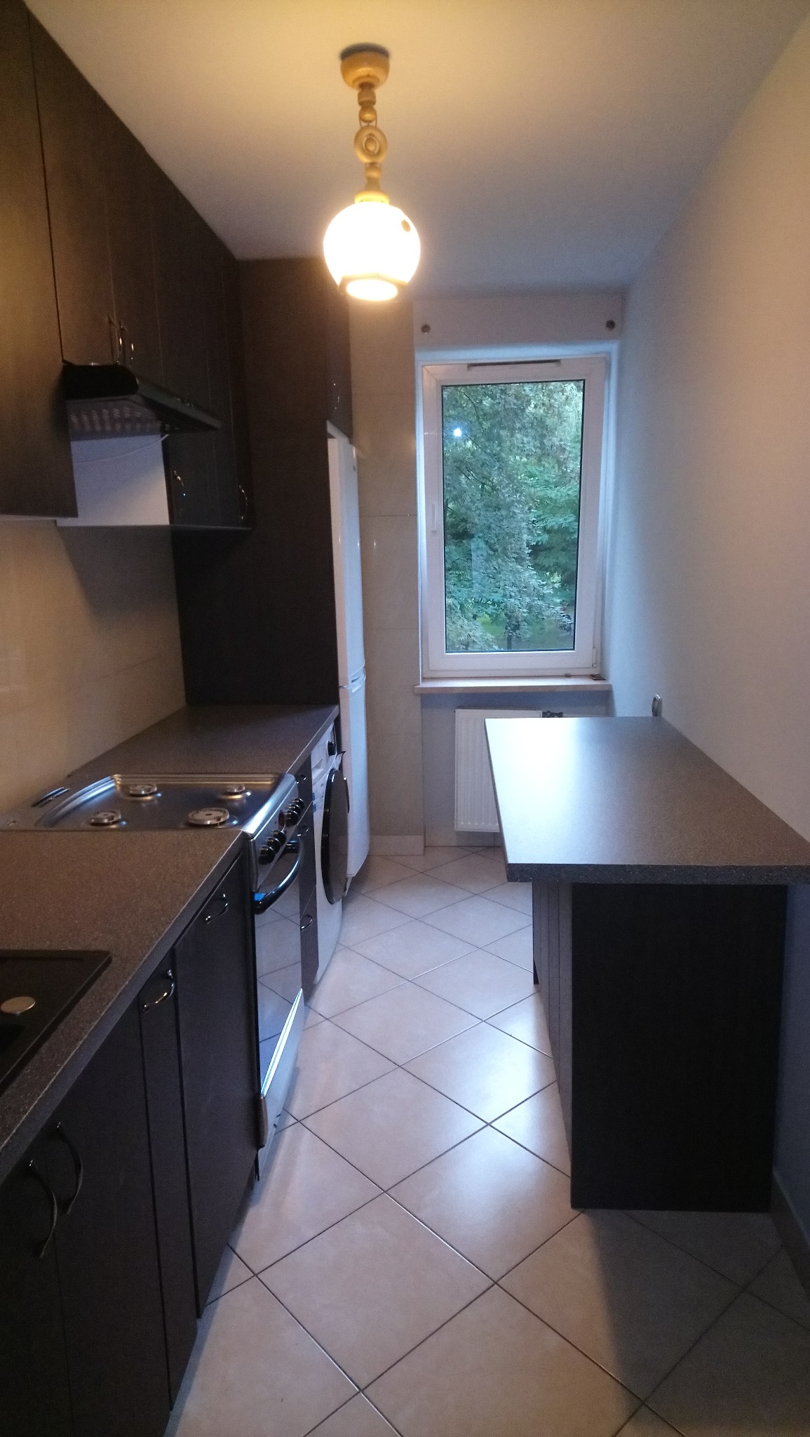 Wąska kuchnia z ciemnymi szafkami, blatem i piekarnikiem, białą lodówką, pralką, i jasną podłogą z płytek w karo, oświetlona lampą sufitową i światłem z okna.