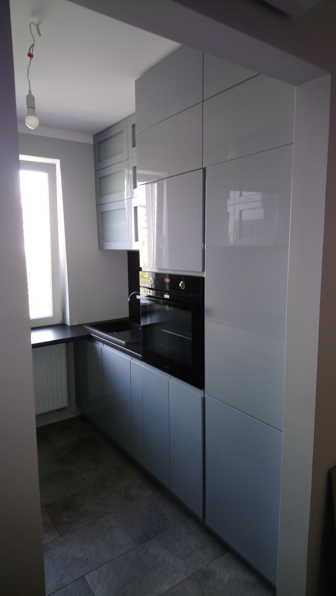 Nowoczesna, szara kuchnia z połyskiem, z zabudowaną czarną piekarnikiem i czarnym blatem, widok z boku, oświetlona naturalnym światłem z okna.