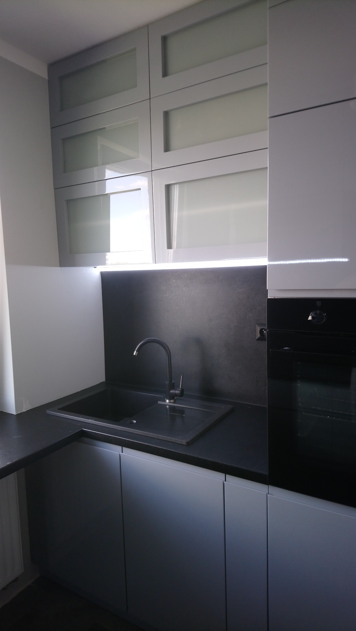 Nowoczesna kuchnia w odcieniach szarości z czarnym blatem i zlewozmywakiem, górne szafki z mlecznym szkłem, oświetlenie LED pod szafkami, piekarnik w zabudowie.