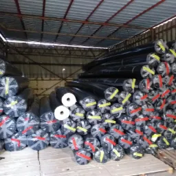 Stosy czarnych, zafoliowanych rolek materiału na drewnianej palecie w magazynie, zabezpieczone taśmami w kolorach żółtym i czerwonym.