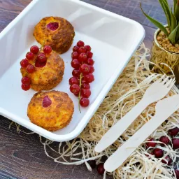 Trzy muffiny z malinami i czerwoną porzeczką w białej, prostokątnej formie obok drewnianych sztućców, dekoracyjnych wiórków i doniczki z zieloną rośliną na ciemnym, drewnianym stole.