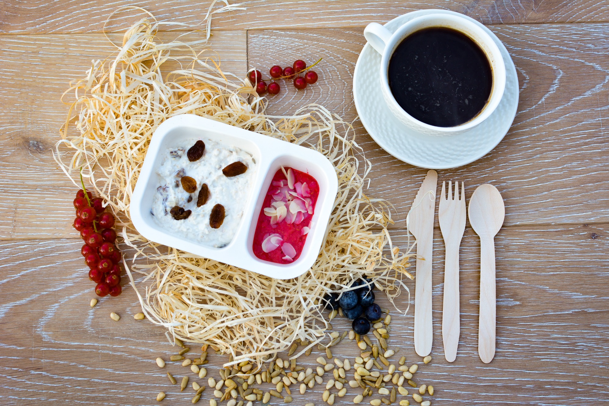 Deser w dwukomorowym pojemniku z owocami i orzechami na drewnianym tle z kawą i sztućcami, całość otoczona dekoracyjną wełną drzewną.