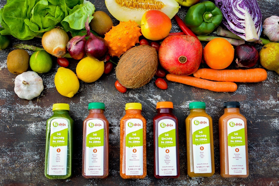 Sześć butelek soków detox 'Fit Dieta' z owocami i warzywami w tle: sałata, kiwi, cebula, kokos, granat, marchew, cytryna, czosnek, jabłko, gruszka, papryka i kapusta.