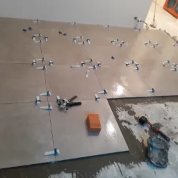 Układanie płytek podłogowych z użyciem systemu poziomowania: szare płytki, krzyżyki dystansowe, poziomica i gumowy młotek widoczne na podłodze w trakcie pracy.