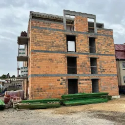 Trzykondygnacyjny budynek w trakcie wznoszenia, ściany z czerwonej cegły dziurawki, widoczne otwory okienne i zbrojenia stropów, na rusztowaniu paleta z cegłami H+H, obok stos zielonych belek...