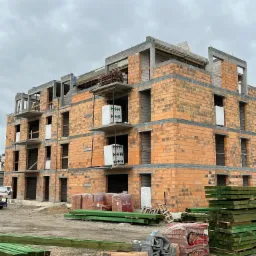 Wielopiętrowy budynek w stanie surowym, wznoszony z czerwonej cegły ceramicznej, widoczne materiały budowlane na placu budowy: palety cegieł, impregnowane drewno konstrukcyjne w kolorze zielonym...