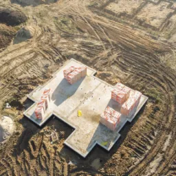 Widok z drona na fundament domu w kształcie litery H, z paletami cegieł na powierzchni, otoczony ziemią z widocznymi śladami opon i wykopami pod przyszłe instalacje.