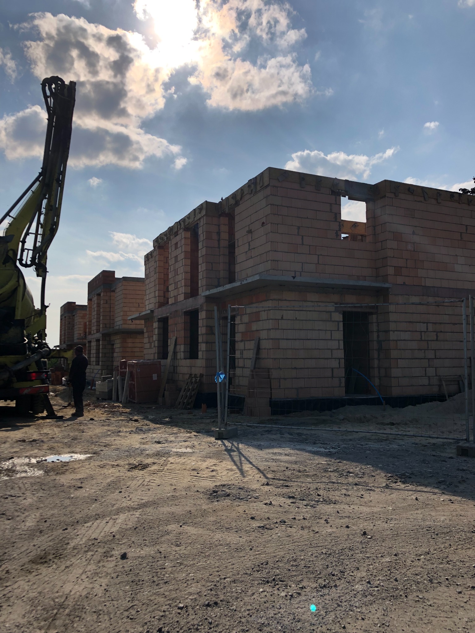 Wznoszone szeregowo budynki z czerwonej cegły, widoczne okna i otwory drzwiowe, na pierwszym planie pompa do betonu i pracownik, w tle pochmurne niebo.