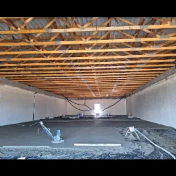 Wnętrze hali z surowym drewnianym stropem, świeżo wylana szara posadzka wyrównywana maszynowo, widoczne narzędzia i materiały budowlane.