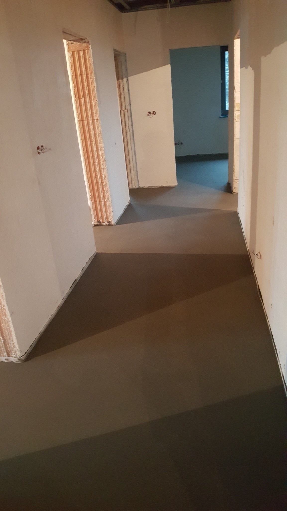 Świeżo wylana, gładka, szara podłoga w surowym wnętrzu z widocznymi pustakami w ościeżnicach drzwiowych. Podłoga rozciąga się przez kilka pomieszczeń, widoczne okno w oddali.
