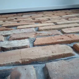 Ułożona ceglana ściana z widoczną szarą fugą, perspektywa z dołu, przy białej listwie przypodłogowej.