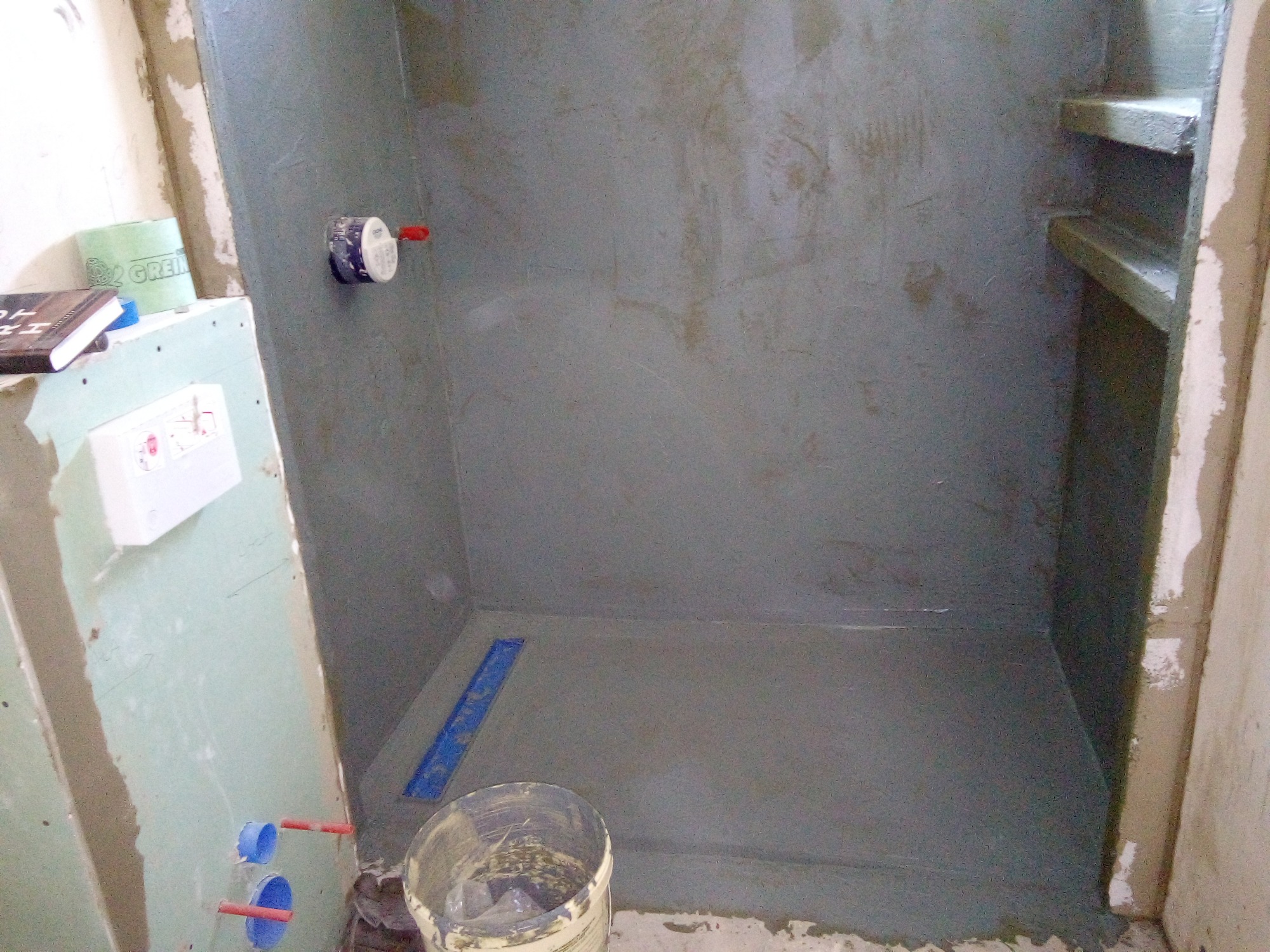 Przygotowanie do hydroizolacji prysznica walk-in w Kolbuszowej: ściany pokryte szarą warstwą izolacyjną, widoczny odpływ liniowy i fragment instalacji wodnej. Na pierwszym planie wiadro...