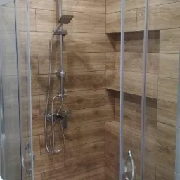 Nowoczesna kabina prysznicowa z deszczownicą i baterią termostatyczną, wykończona płytkami imitującymi drewno, w tle widoczna pralka i umywalka.