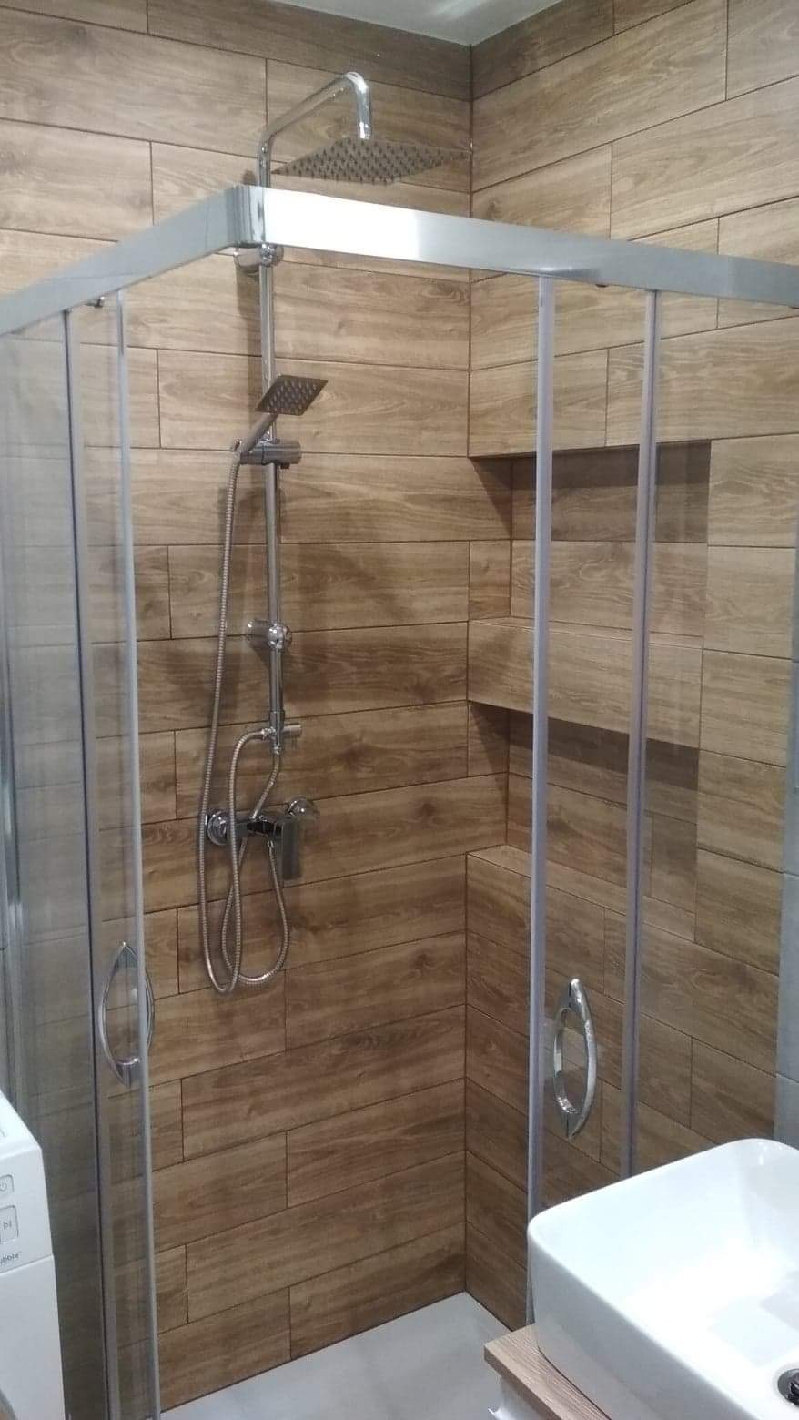 Nowoczesna kabina prysznicowa z deszczownicą i baterią termostatyczną, wykończona płytkami imitującymi drewno, w tle widoczna pralka i umywalka.