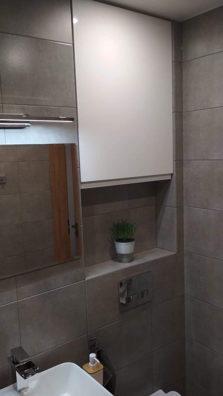 Nowoczesna łazienka z szarymi płytkami, białą szafką nad toaletą, umywalką z chromowaną baterią i rośliną doniczkową w niszy.