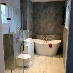 Nowoczesna łazienka z wolnostojącą wanną, umywalką nablatową i podwieszaną toaletą, wykończona szarymi płytkami imitującymi kamień i jasną podłogą z efektem drewna, z widoczną instalacją...