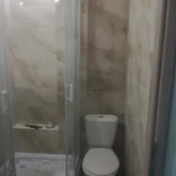 Ustawiona pod kątem kabina prysznicowa z półką wewnątrz, obok toaleta, całość w łazience wyłożonej marmurkowymi płytkami.