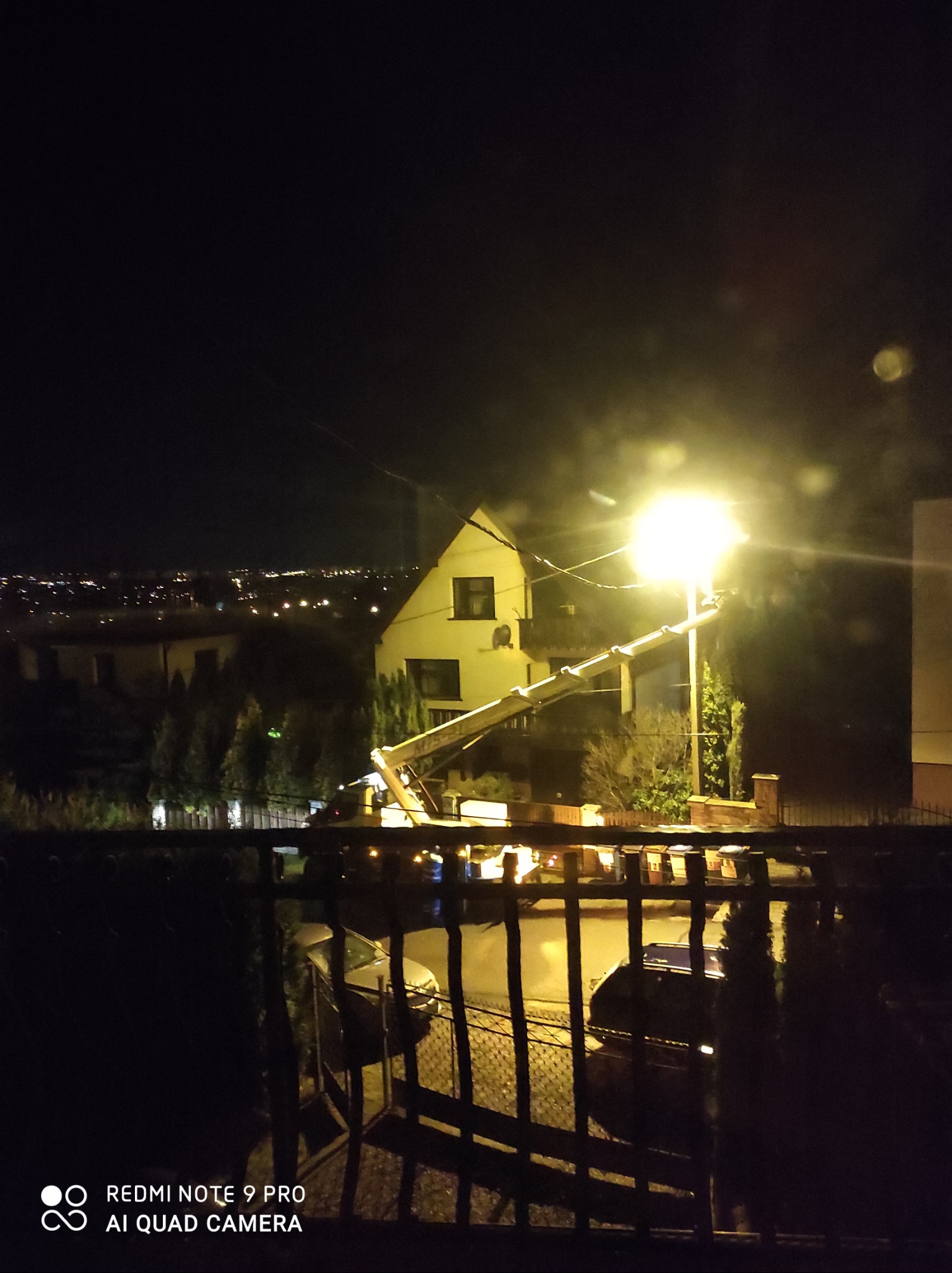 Nocny widok na dom z podnośnikiem koszowym oświetlonym jasnym światłem, prawdopodobnie podczas prac na wysokościach, widoczne zabudowania w oddali.