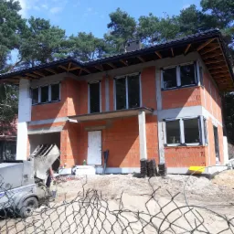 Budowa domu jednorodzinnego z czerwonej cegły, widoczna maszyna do jastrychu na zewnątrz, częściowo ogrodzona siatką.