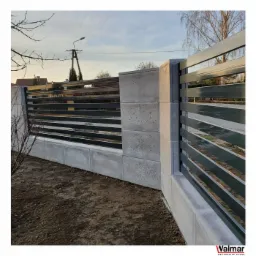 Nowoczesne ogrodzenie panelowe z betonowymi słupkami i podmurówką, poziome przęsła w kolorze grafitowym, widoczny fragment ogrodu i słup energetyczny w tle.