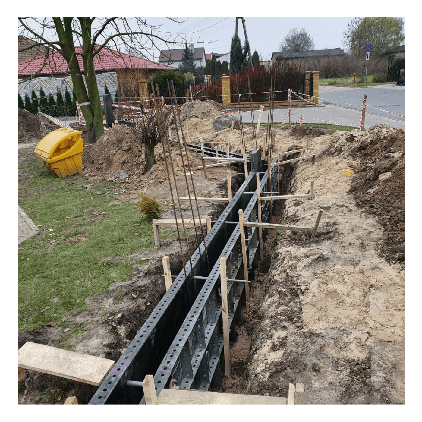 Przygotowanie wykopu i szalunku pod fundament ogrodzenia panelowego na działce z widocznym żółtym pojemnikiem na śmieci i domami w tle.