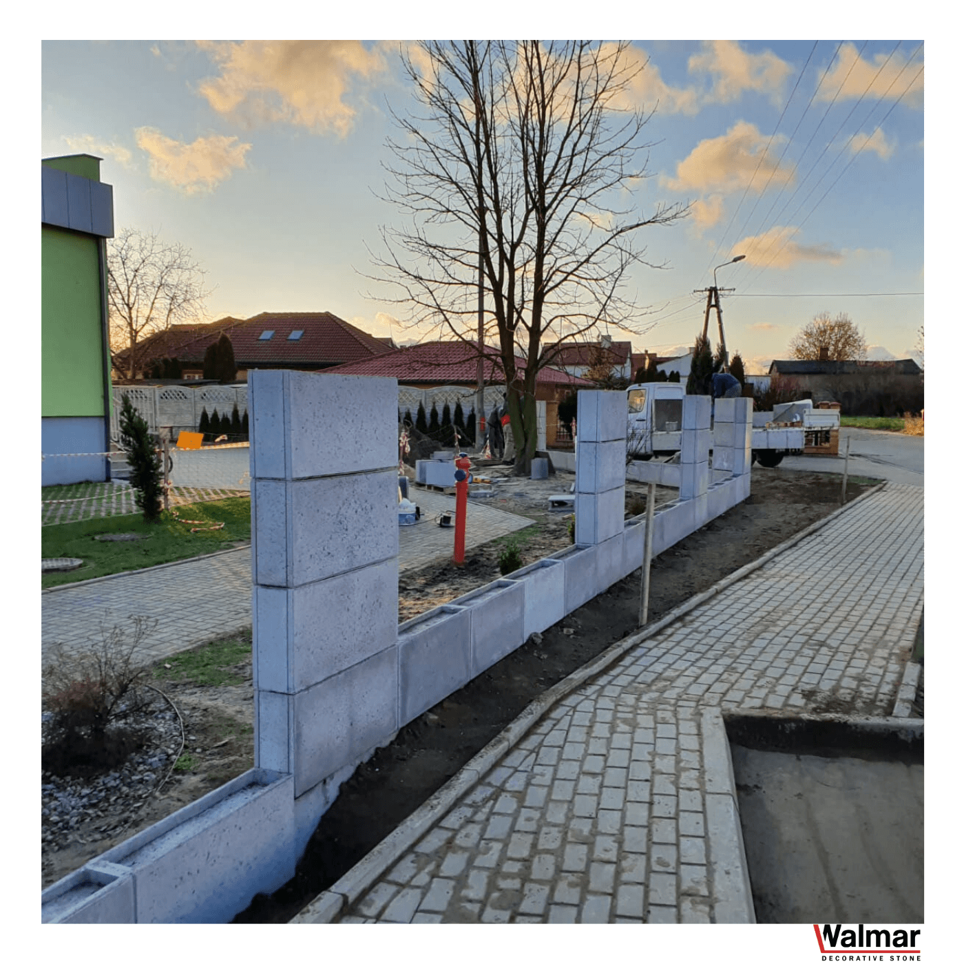 Budowa ogrodzenia z szarych betonowych bloczków przy chodniku z kostki, widoczne elementy konstrukcyjne i narzędzia, w tle domy jednorodzinne i zachodzące słońce.